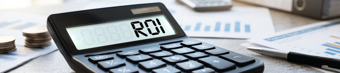 A close-up of a black calculator on a desk displaying "ROI" on its screen, surrounded by stacks of coins, a pen, and financial charts in the background.