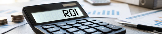 A close-up of a black calculator on a desk displaying "ROI" on its screen, surrounded by stacks of coins, a pen, and financial charts in the background.