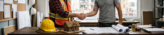 Two men shaking hands over a desk with blueprints, a yellow hard hat, and a small model house inside a construction office.